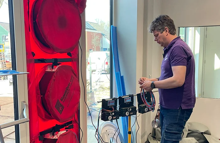 Man in een paarse polo bedient meetapparatuur naast een deur die is afgedicht met een rood blowerdoor-systeem tijdens een luchtdichtheidstest.